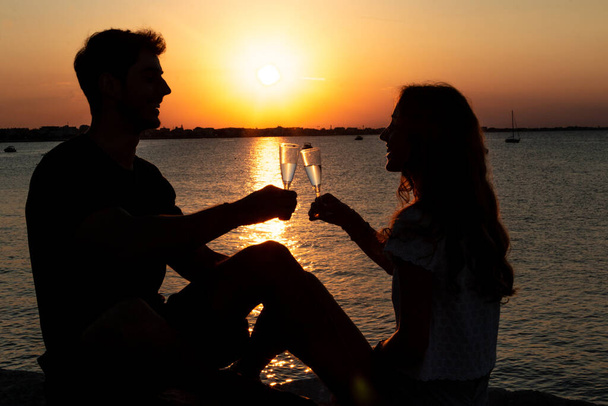 Verliebtes Paar am Strand von Sonnenuntergang - zwei junge Liebende stoßen im Sonnenuntergang an - Foto, Bild
