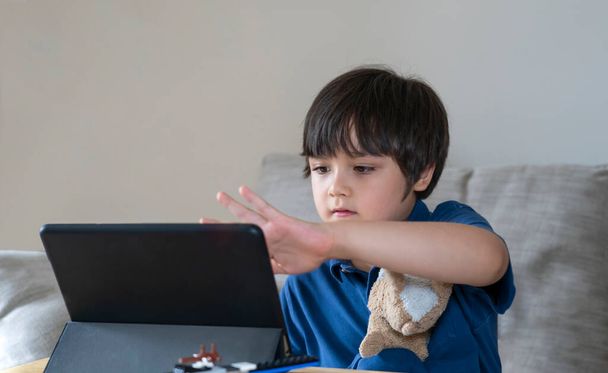 School Kid za pomocą tabletu do pracy domowej, Dziecko patrząc na cyfrowy tablet z myślącą twarzą, Młody chłopiec oglądając kreskówkę na dotyku pad, Nowy normalny styl życia z uczenia się online, Edukacja na odległość - Zdjęcie, obraz