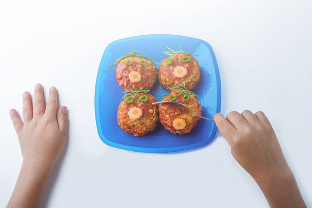 Child food. Funny food. Plate with buckwheat cutlets or meatballs in the form of funny faces decorated with greens. Children's or kids menu. Healthy eating. Kid's hands hold the fork - Фото, изображение