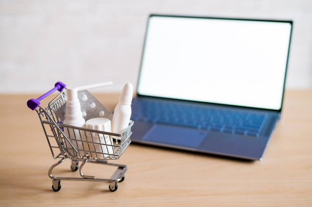 Laptop with blank white screen and miniature shopping cart full of medicines. Online pharmacy concept. Website for buying tablets. - Фото, зображення