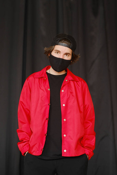 Portrait of a young man 25-30 years old in a black protective mask, black cap and red jacket.  - Fotoğraf, Görsel