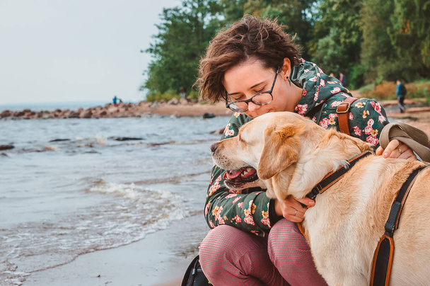 Una ragazza bacia un cane sulla testa sullo sfondo del mare. Passeggiata con un Labrador fulvo su una spiaggia sabbiosa - Foto, immagini