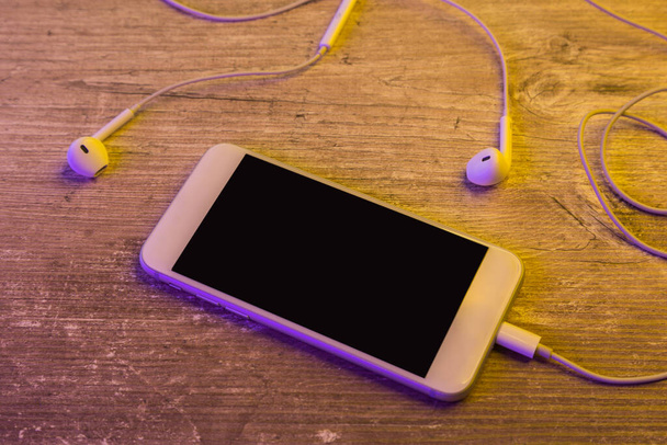 älypuhelin, jossa on valkoiset kuulokkeet puupöydällä neonvalolla, moderni - Valokuva, kuva