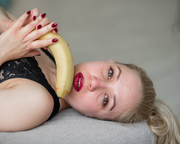 The blonde in black lace underwear erotically eats a banana. Attractive woman with sensual red lips sexually sucks and licks a banana. Oral pleasure. - Photo, image