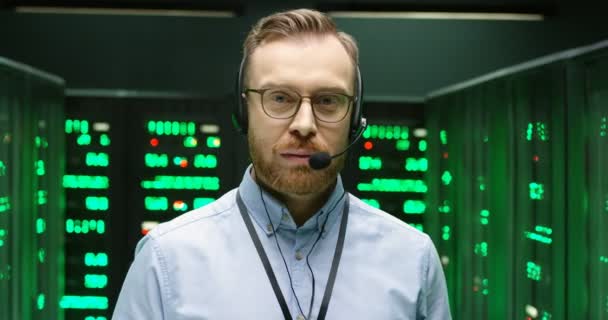 Portré kaukázusi jóképű férfi szemüvegben és fülhallgató áll az adatbázisban szoba szerverek és nézte a kamerába. A férfi biztonsági dolgozó közelsége az adatközpontban működő feldolgozókkal. - Felvétel, videó
