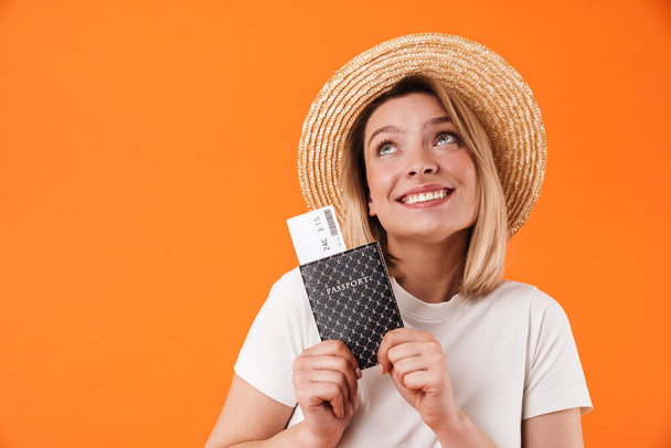Image of happy woman in straw hat smiling and holding passport with tickets isolated over orange background - Фото, изображение