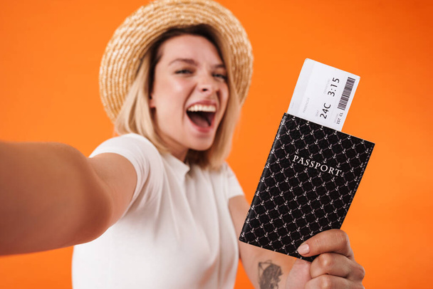Afbeelding van opgewonden vrouw met paspoort met tickets en het nemen van selfie foto geïsoleerd over oranje achtergrond - Foto, afbeelding