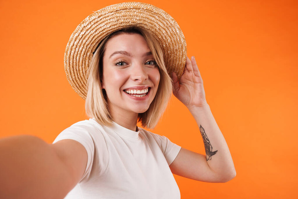 Image of cheerful young woman in straw hat smiling and taking selfie photo isolated over orange background - Fotografie, Obrázek