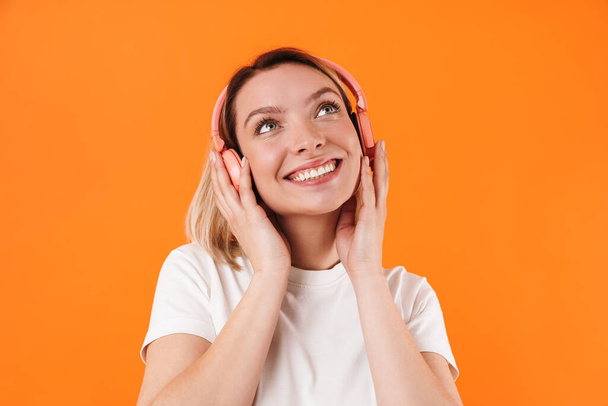 Portrait of lovely cheerful young blonde woman wearing casual clothes standing isolated over orange background, listening to music with wireless headphones - Photo, Image