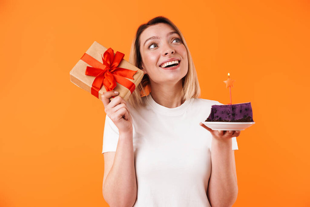 Portrait of lovely cheerful young blonde woman wearing casual clothes standing isolated over orange background, showing present box, holding birthday cake - Fotografie, Obrázek