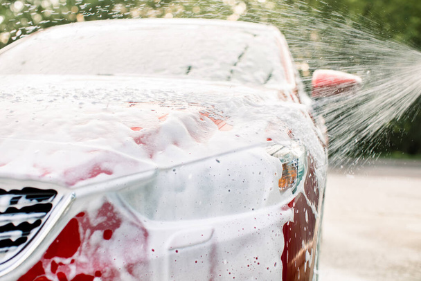 Close up horizontal shot of red car with cleaning foam, washed at car wash service outdoors with high pressure sprayer. Clean car concept. Car wash outdoor - Foto, Imagem