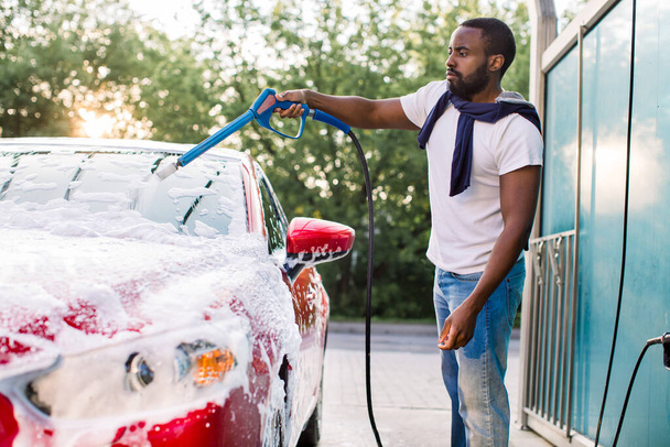 Portrait of handsome bearded young African man washing his red car with foam at at self service carwash outdoors. Clean car concept. Car wash - Foto, Imagem