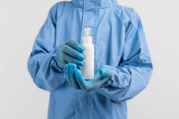 doctor in a protective medical suit against coronavirus, wearing latex gloves, holds a jar of antiseptic disinfectant on an isolated white background. Coronavirus, quarantine - Photo, image