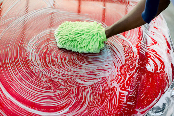 Closeup cropped image of hand of African man with green sponge washing his red car hood at a self-serve car wash outdoors. Hood of red luxury car covered by soap. - Foto, imagen