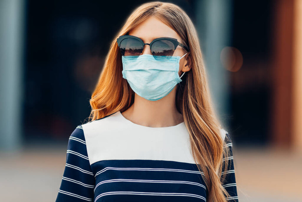 young attractive girl in sunglasses and a medical protective mask on her face, standing in the city against the background of a building. Quarantine, coronavirus, - Photo, Image