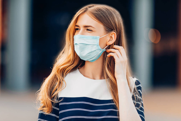 attractive young woman in a medical protective mask on her face, standing in the city against the background of a building. Quarantine, coronavirus, - Фото, изображение