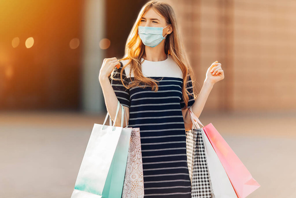 stylish young woman in a medical protective mask on her face, carrying shopping bags, leaving the shopping center after shopping. Quarantine, coronavirus - Photo, image