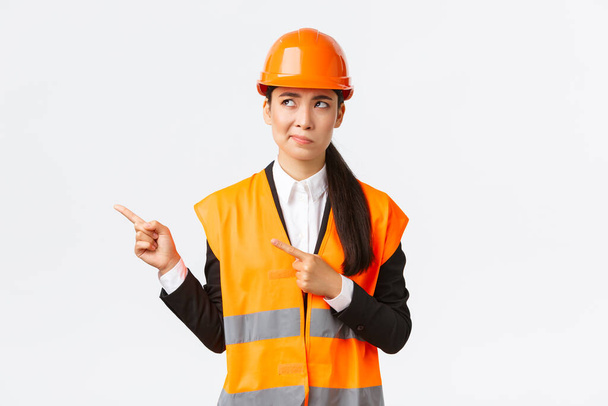 Building, construction and industrial concept. Indecisive asian female architect being unsure, wearing reflective clothing and helmer, smirk as pointing and looking upper left corner puzzled - Photo, Image