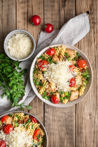 Chicken pasta salad with roasted and sun-dried tomatoes, topped with green onion slices and grated cheese on wooden background - 写真・画像