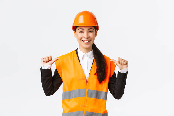 Onnistunut ylpeä hymyilevä aasialainen nainen rakennuspäällikkö, insinööri turvakypärä osoittaa itseään, keuliminen. Arkkitehti suosittelee henkilökohtaista apua, kehuskelu saavutuksia - Valokuva, kuva