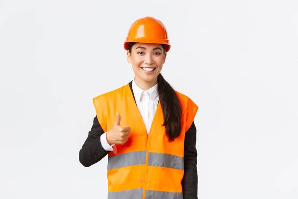Magabiztos elégedett ázsiai női építőipari vezető a biztonsági sisak, fényvisszaverő kabát, mutatja hüvelykujj-up jóváhagyása, támogatás és hasonló munka az alkalmazottak, álló fehér háttér - Fotó, kép