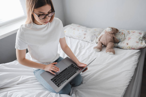 Young smiling woman uses laptop at home on the bed. Communication, video call, remote work. High quality photo - Фото, изображение