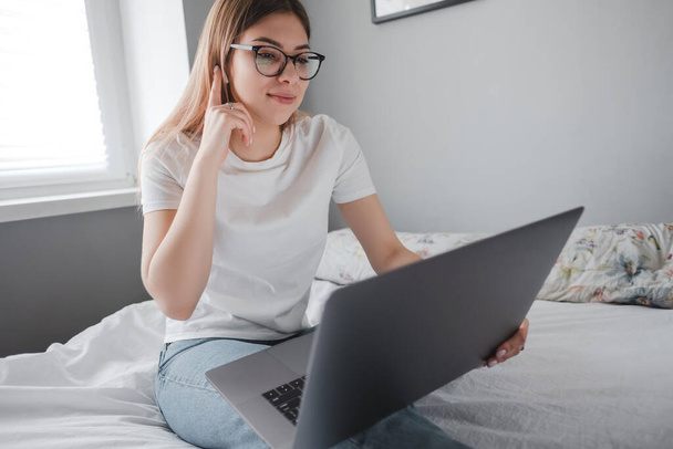 Young smiling woman uses laptop at home on the bed. Communication, video call, remote work. High quality photo - Photo, Image