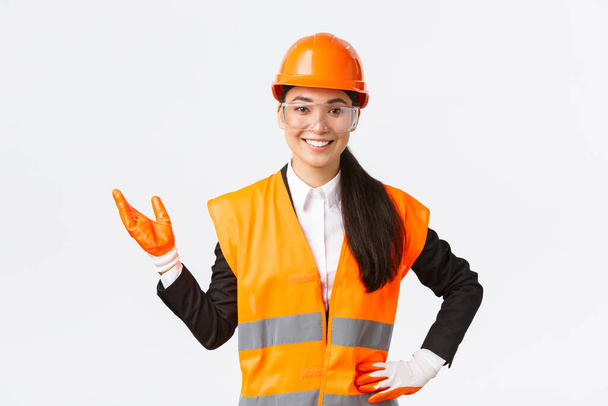 Smiling cheerful asian female construction manager introduce her interprise for investors, wearing safety glasses, helmet and gloves pointing hand left and looking proud, white background - Photo, Image