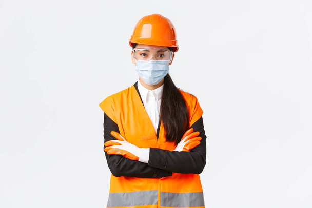 Covid-19 safety protocol at enterpise, construction and preventing virus concept. Confident asian female chief engineer looking how workers follow protocol during coronavirus, wear face mask - Photo, Image