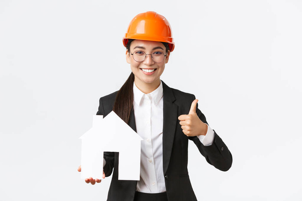 Portrait of young asian female construction engineer, architect in safety helmet and business suit holding home maket and show thumbs-up, promoting company service of building houses - Photo, Image