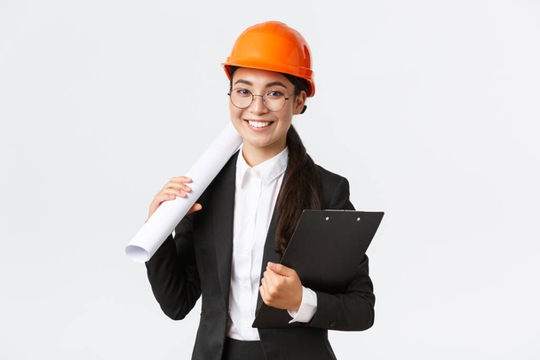 Úspěšná asijská architektka v obleku a helmě s modrotisky a poznámkovými bloky, inspektorka při pohledu na stavby, s úsměvem na kameru, bílé pozadí - Fotografie, Obrázek