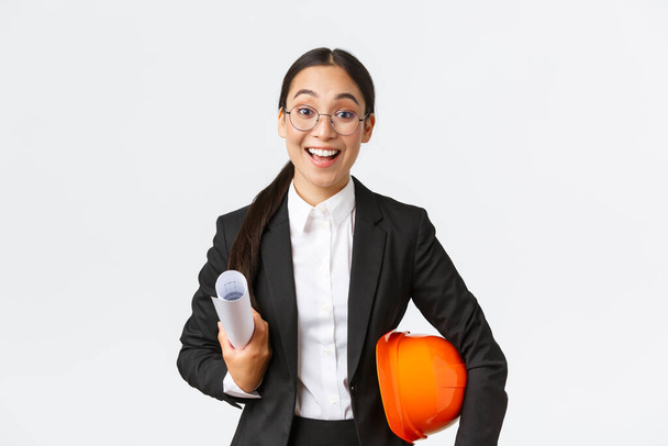 Talentovaná vzrušená asijská architektka zahajující stavební práce, držící modrotisky a bezpečnostní přilbu, v obleku, zasedací inženýři na staveništi, šťastně se usmívající, bílé pozadí - Fotografie, Obrázek