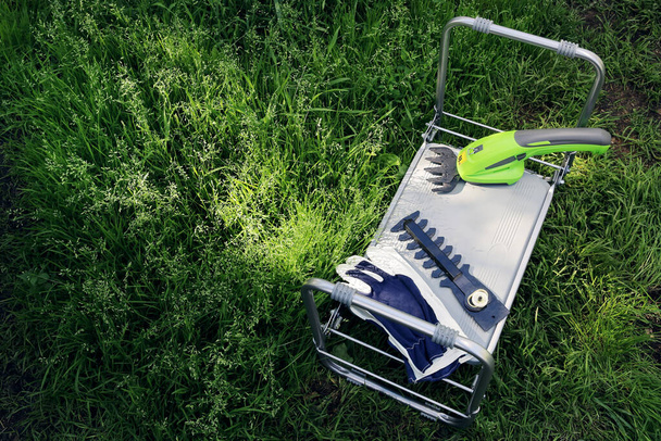 Переносные электрические садовые треугольники с кожаной головкой и кожаными перчатками лежат на раскладном рабочем стуле в зеленой траве солнечным летним днем - Фото, изображение