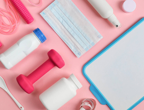 Sports items: dumbbell, measuring tape, protein drink, skipping rope, disinfectant, mask and writing board lie on a pink background, top view close up. - Photo, Image