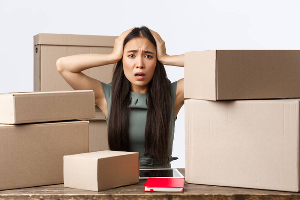 Small business owners and e-commerce concept. Worried asian businesswoman panicking, cant process all orders from her online shop, grab head anxious as sitting with lots of boxes ready for shipping - Foto, imagen