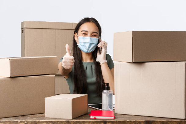 Small business owners, e-commerce and covid-19 preventing virus concept. Happy asian businesswoman in medical mask and gloves answering customer calls, ensure buying is safe, packing products - Photo, Image
