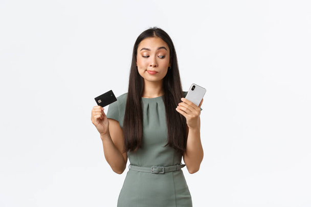 Koncepcja małych przedsiębiorstw, kobiet przedsiębiorców. niezdecydowana sceptyczna azjatycka kobieta próbuje nowej aplikacji bankowej i patrząc wątpliwe na ekranie smartfona pokazując kartę kredytową - Zdjęcie, obraz