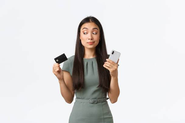 Small business owners, women entrepreneurs concept. Hesitant skeptical asian woman trying new banking application and looking doubtful at credit card while holding mobile phone, white background - Foto, imagen