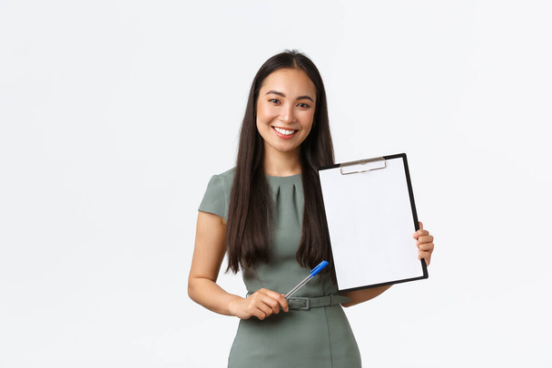 Gülümseyen Asyalı iş kadını kağıtlarını, açıklama belgelerini gösteriyor, boş panoyu gösteriyor ve kalem kullanarak bilgiyi tanımlıyor, beyaz arka planda duruyor. Çalışanların sunumu - Fotoğraf, Görsel