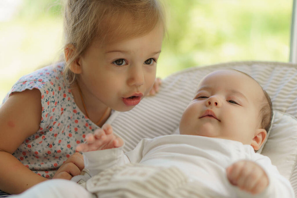 Roztomilá holčička líbající jeho novorozenou sestru. Dítě batole potkává nově narozeného sourozence. Dítě spí v bílém vyhazovači pod přikrývkou. Děti si hrají a sbližují se. Děti s malým věkovým rozdílem. - Fotografie, Obrázek