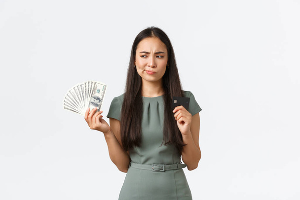 Kleinunternehmer, Unternehmerinnen. Unentschlossene asiatische Frau denkt, während sie Kreditkarte und Bargeld in der Hand hält, ratlos, wie sie den Kauf bezahlen soll, steht nachdenklich vor weißem Hintergrund - Foto, Bild