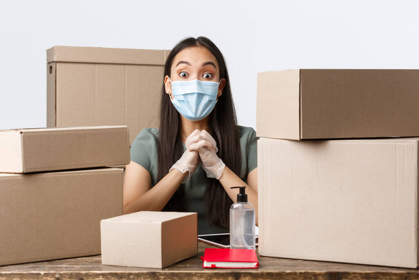 Small business owners, e-commerce and covid-19 preventing virus concept. Excited and amazed asian woman in medical mask and gloves selling online, packing customer order in boxes, use hand sanitizer - Fotó, kép