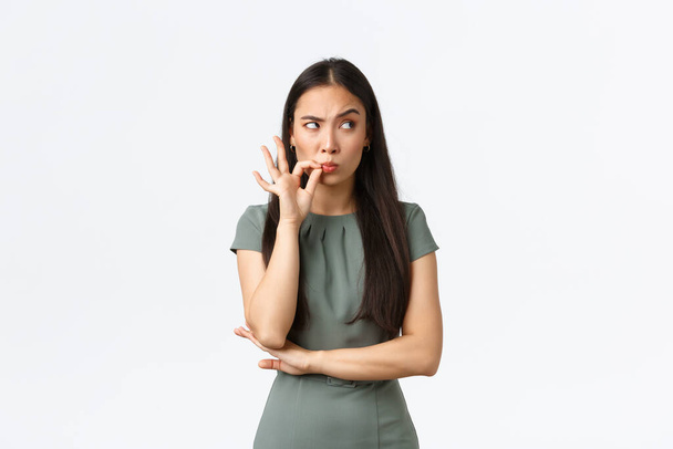 Small business owners, women entrepreneurs concept. Suspicious asian woman looking with disbelief upper right corner, feeling doubtful, zipping mouth, seal lips as making promise not speak - Photo, Image