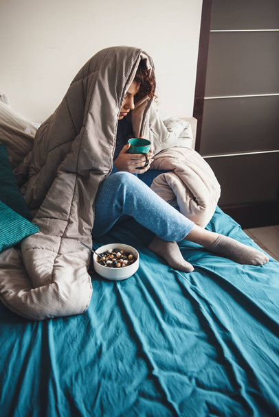 Γοητευτική κυρία με σγουρά μαλλιά που πίνει γάλα και τρώει δημητριακά στο κρεβάτι καλυμμένη με πάπλωμα - Φωτογραφία, εικόνα