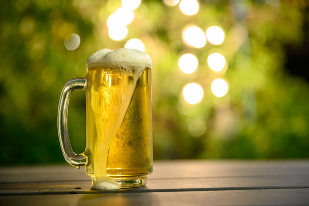Szklanka zimnego piwa z pianką na szczycie Umieszczone na stole w ogrodzie, tło jest zielonym drzewem i ma piękny bokeh o zmierzchu. - Zdjęcie, obraz