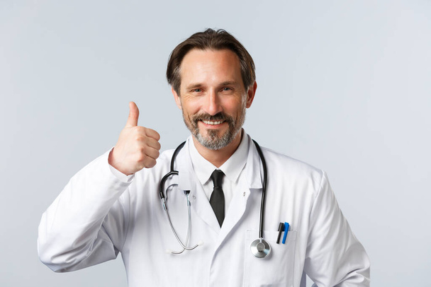 Covid-19, preventing virus, healthcare workers and vaccination concept. Close-up of satisfied and pleased doctor in white coat, show thumb-up in approval, recommend medical product or clinic service - Φωτογραφία, εικόνα