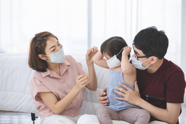 Азиатская семья в медицинских масках смотрит в камеру, стоя дома в гостиной, чтобы предотвратить PM2.5 пыль, смог, загрязнение воздуха и COVID-19. Концепция здравоохранения. - Фото, изображение
