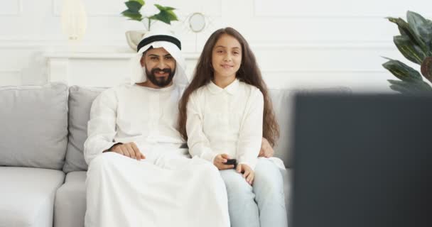 Arab young handsome father sitting on couch with small teen cute daughter and watching TV. At home. Little teenage girl changing channels with remote control and watch television with dad in cozy room - Video
