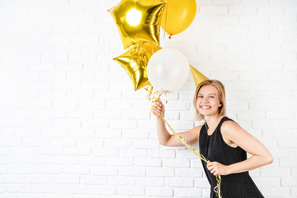 Birthday party. Young blond smiling woman wearing birthday hat holding golden balloons celebrating birthday with copy space - 写真・画像