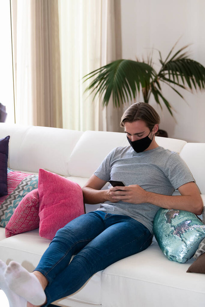Hezký mladík s obličejovou maskou, který kontroluje telefon, zatímco sedí na pohovce na rozostřeném pozadí. Relaxační a komunikační koncepce. - Fotografie, Obrázek